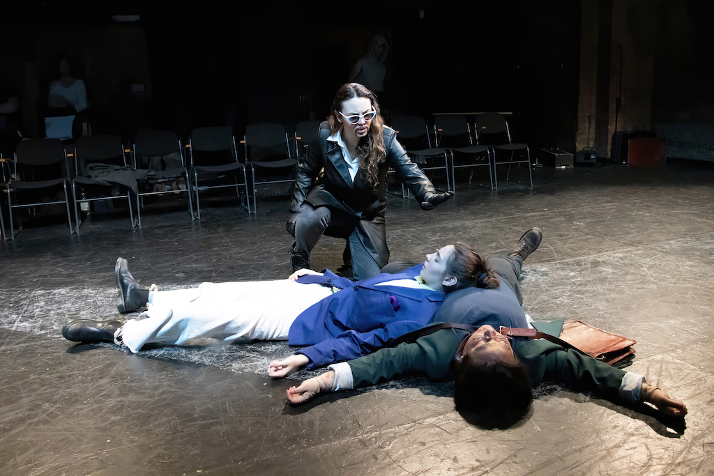 Two actors lie with eyes closed on top of one another on stage while a third actor wearing sunglasses and a goatee crouches over them in mid-performance.
